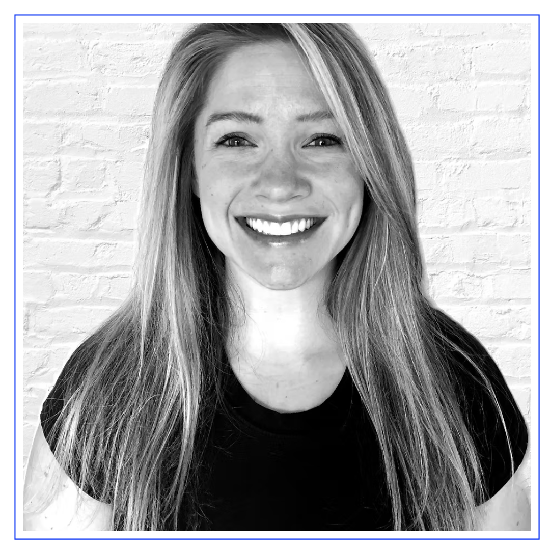 A woman with long hair is smiling at the camera, wearing a black shirt. The image is in black and white, with a white brick wall in the background.