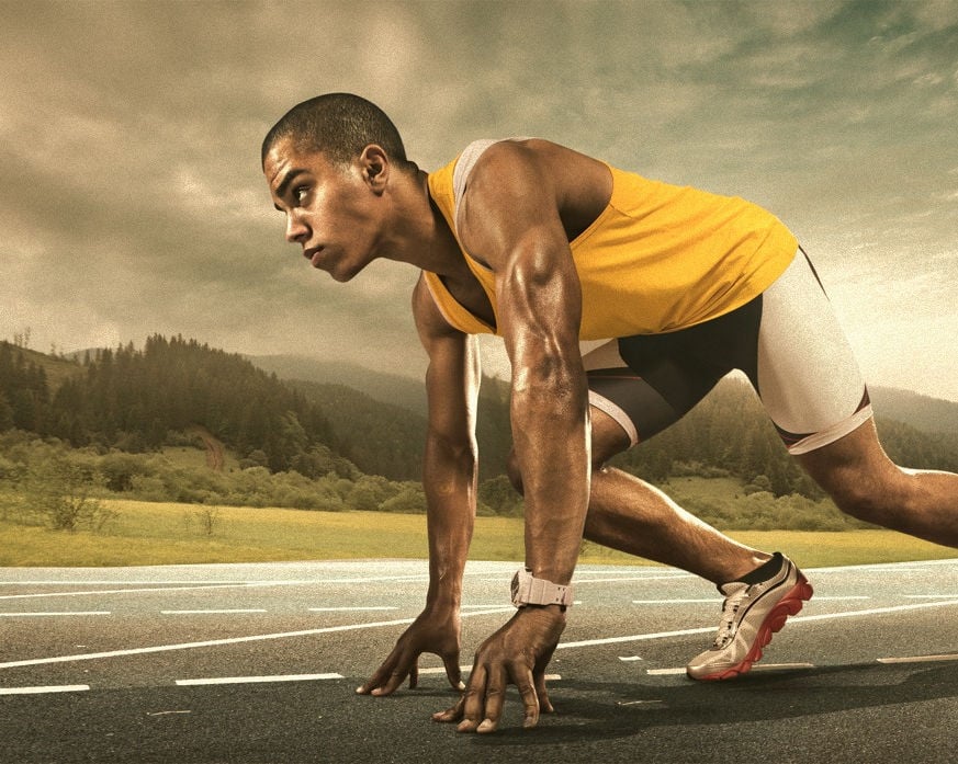 A male athlete is crouched in a starting position on a running track, preparing to race. The background shows a scenic view of mountains and fields.